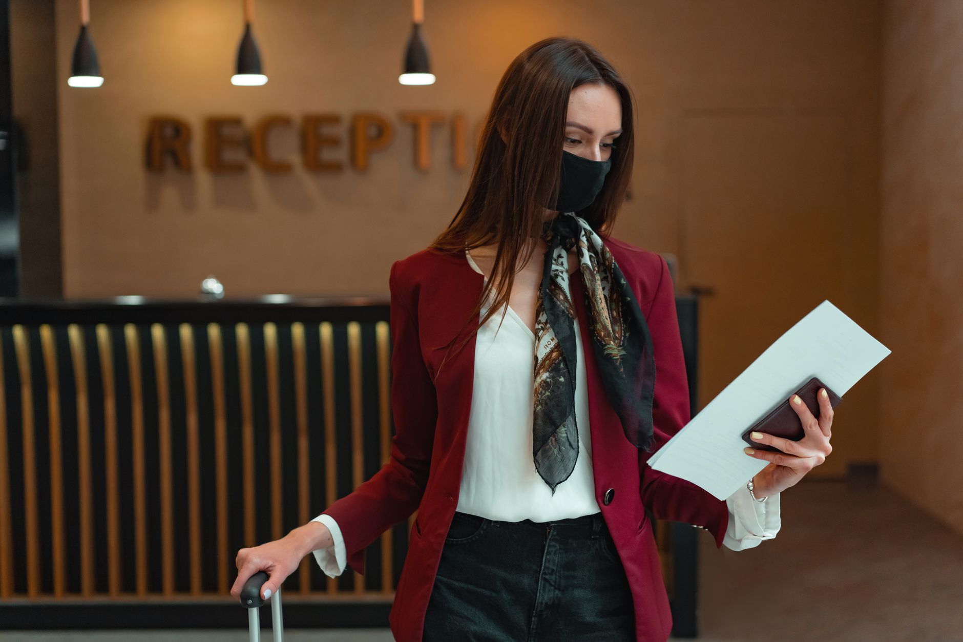 Kobieta z bagażem i dokumentami przy recepcji hotelowej w maseczce